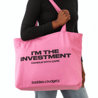 Black woman holding a pink canvas tote that reads “I’M THE INVESTMENT – HANDLE WITH CARE” by Baddies & Budgets
